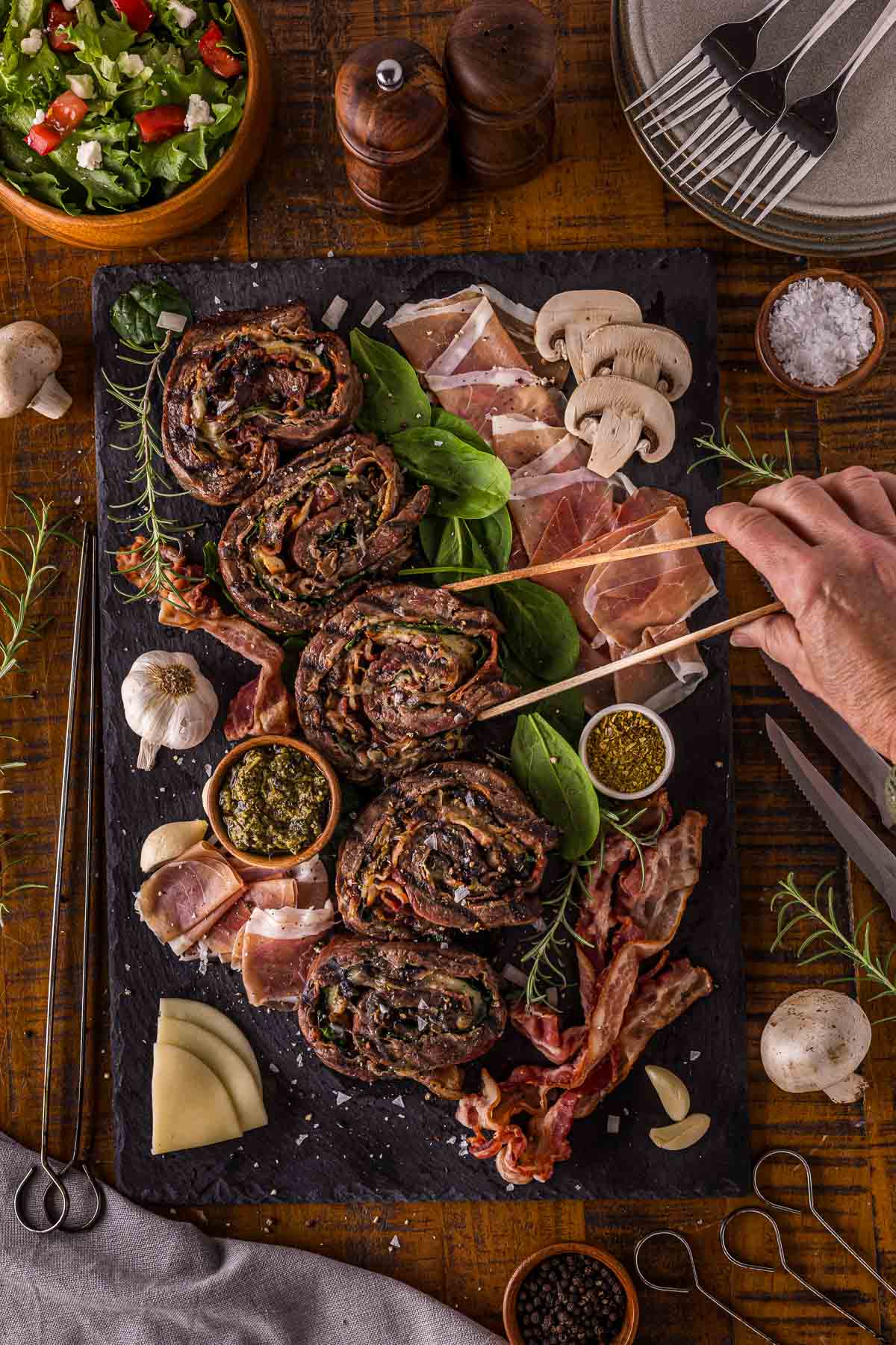 A hand slices rolled grilled beef stuffed with spinach on a slate board, surrounded by prosciutto, bacon, cheese, mushrooms, pesto, garlic, fresh salad, and seasonings.