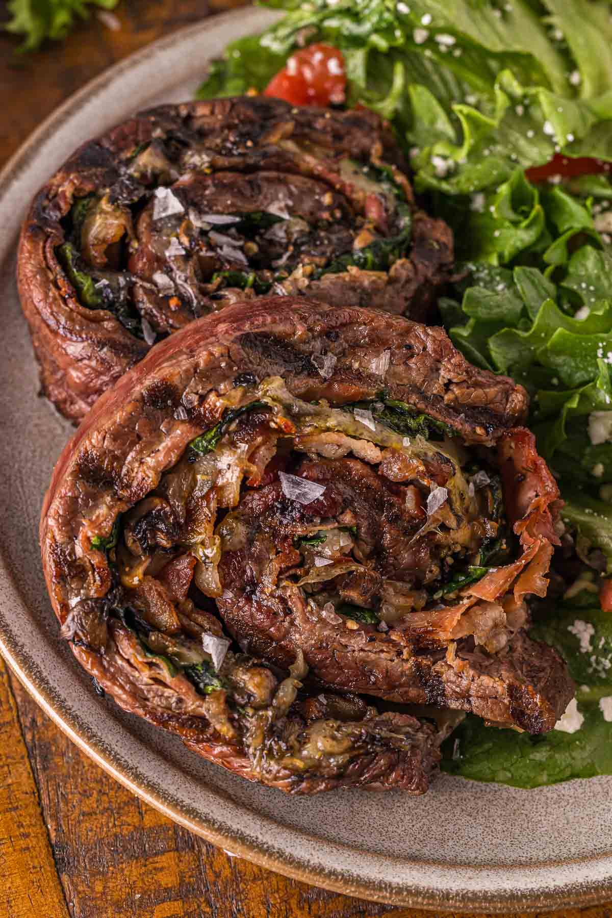 Two flank steak pinwheels served on a plate with a green leafy salad.