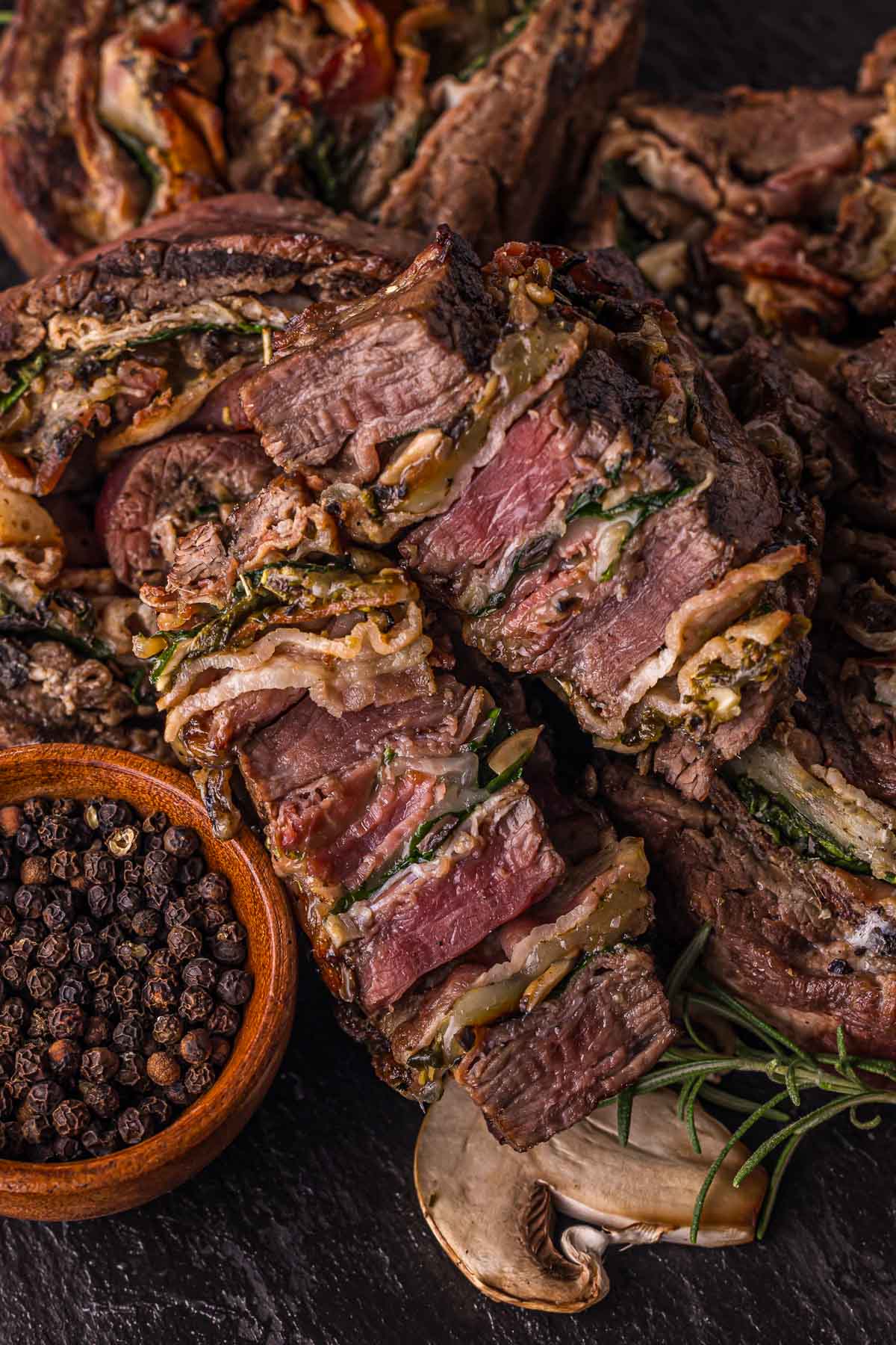 Sliced stuffed flank steak pinwheels with layers of mushrooms, spinach, and cheese, served on a dark surface with black pepper and fresh rosemary.