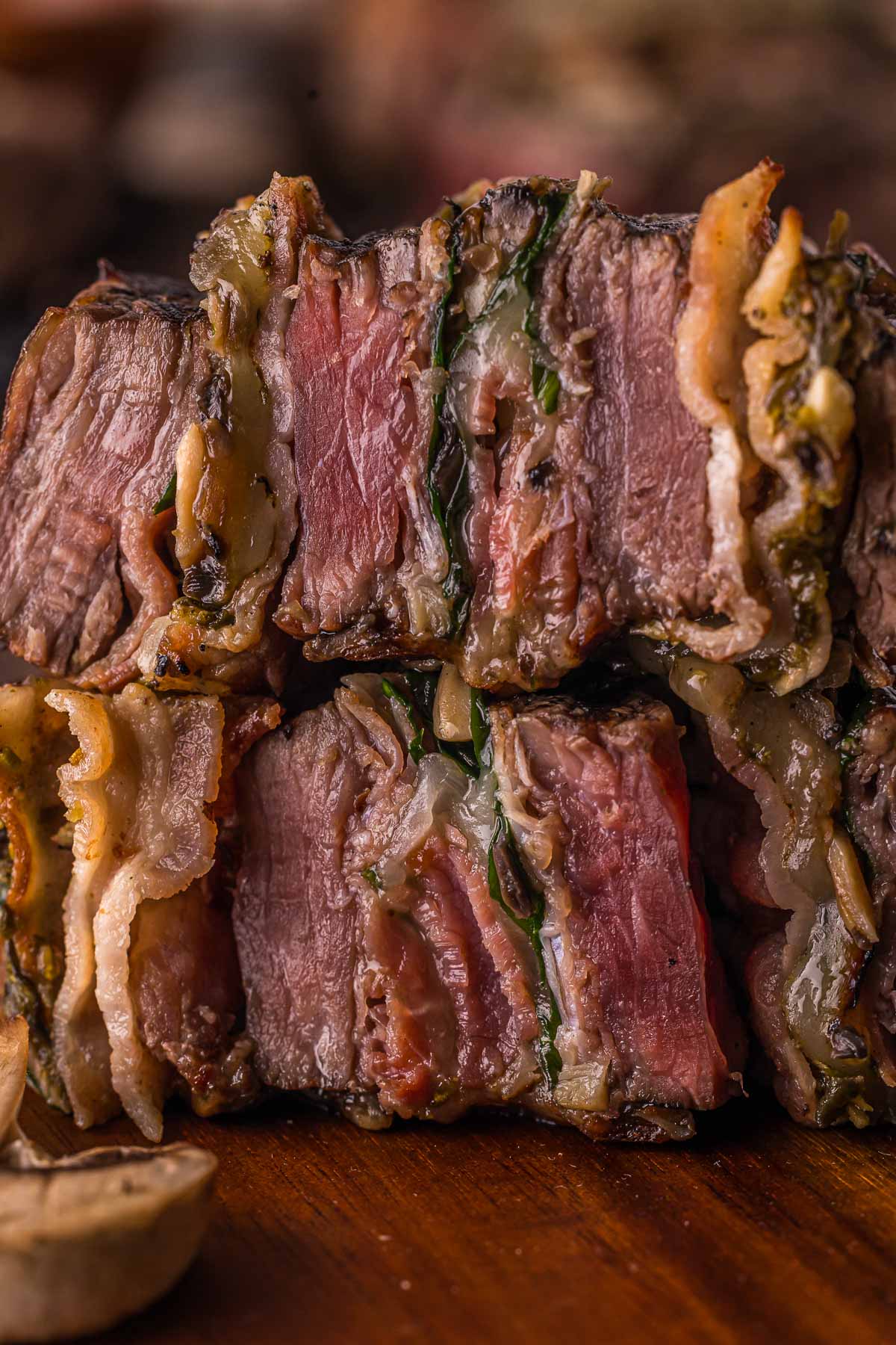 Close-up of two thick slices of medium-rare flank steak pinwheels with crispy bacon and filling, stacked on a wooden surface.
