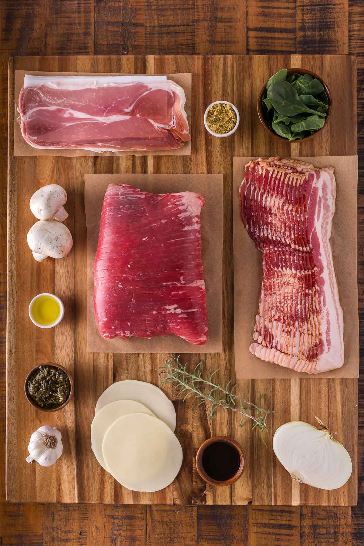 A wooden board displays raw flank steak, bacon strips, prosciutto, mushrooms, spinach, provolone cheese, seasonings, olive oil, garlic, and a sprig of rosemary.
