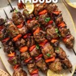Overhead view of grilled lamb kabobs with red peppers and onions, garnished with herbs, served with lemon wedges and flatbread on a plate.