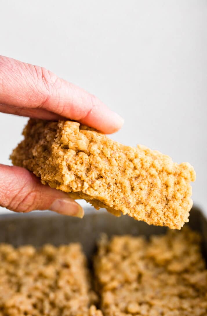 A hand holding a soft and chewy homemade rice crispy bar over a pan of sliced bars.