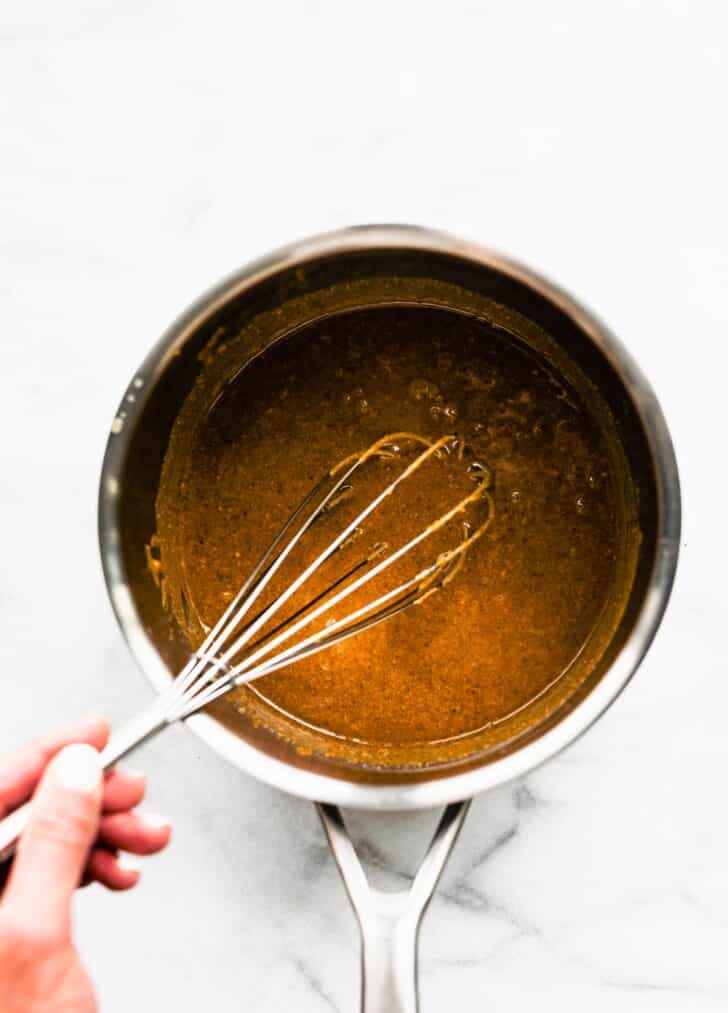 1. A homemade maple and nut butter mixture being whisked in a saucepan on a marble countertop.