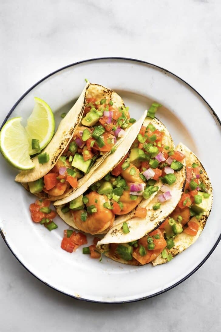 3 shrimp tacos on a white plate topped with avocado and chopped onion and a lime wedge on the plate