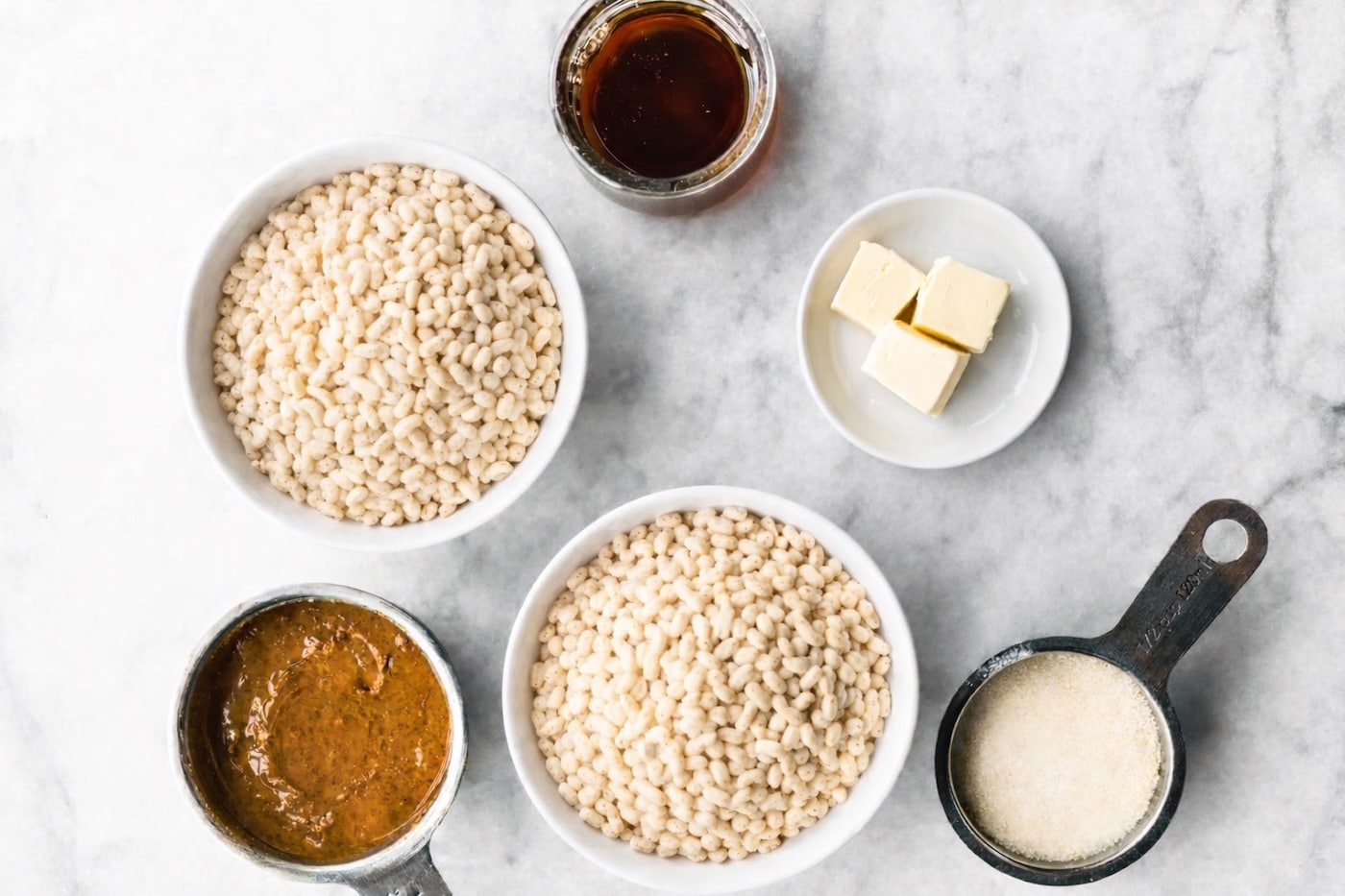 Ingredients for homemade rice crispy bars arranged on a marble surface, including crispy rice cereal, maple syrup, butter, and a nut butter mixture.