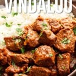 A bowl of lamb vindaloo served with white rice, garnished with herbs; text reads "How to Make Lamb Vindaloo" and includes a website for the recipe.