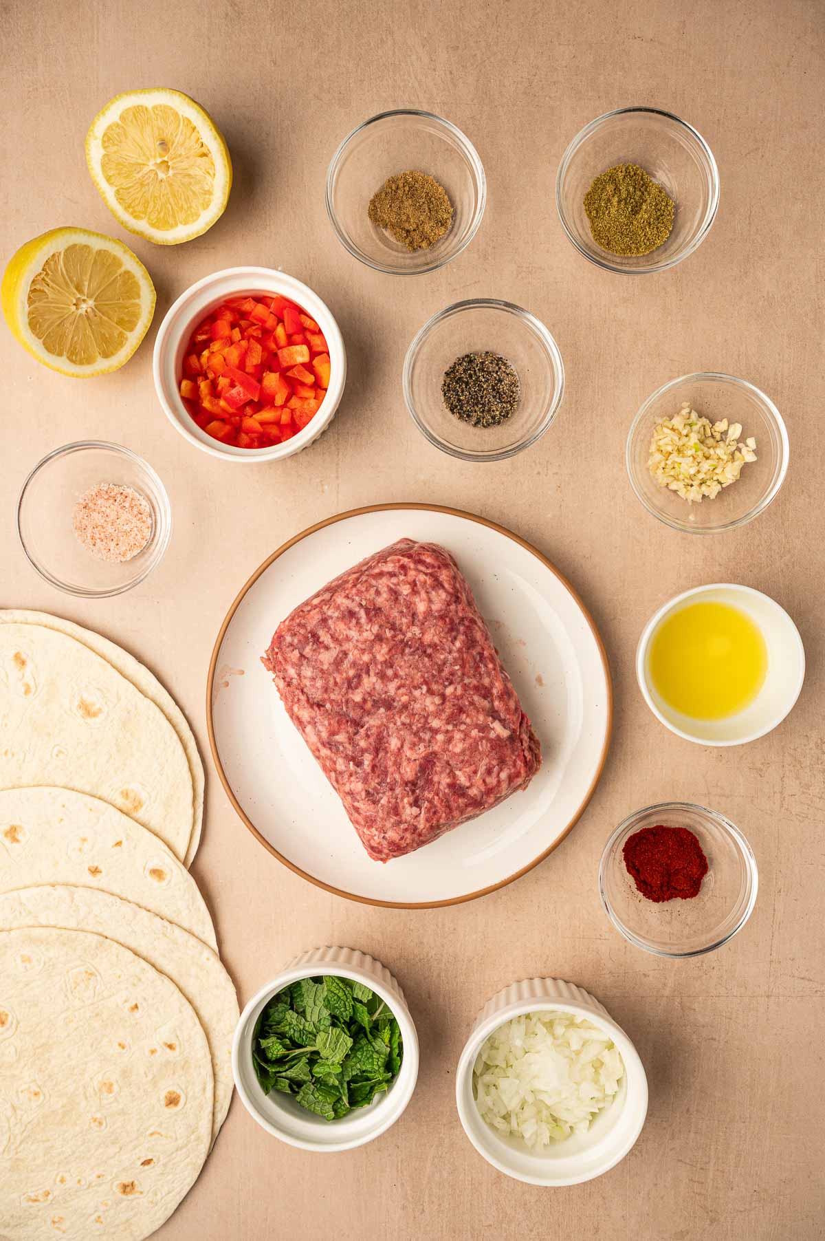Flat lay of ingredients including ground lamb, tortillas, chopped vegetables, lemon halves, spices, olive oil, and herbs arranged on a beige surface.
