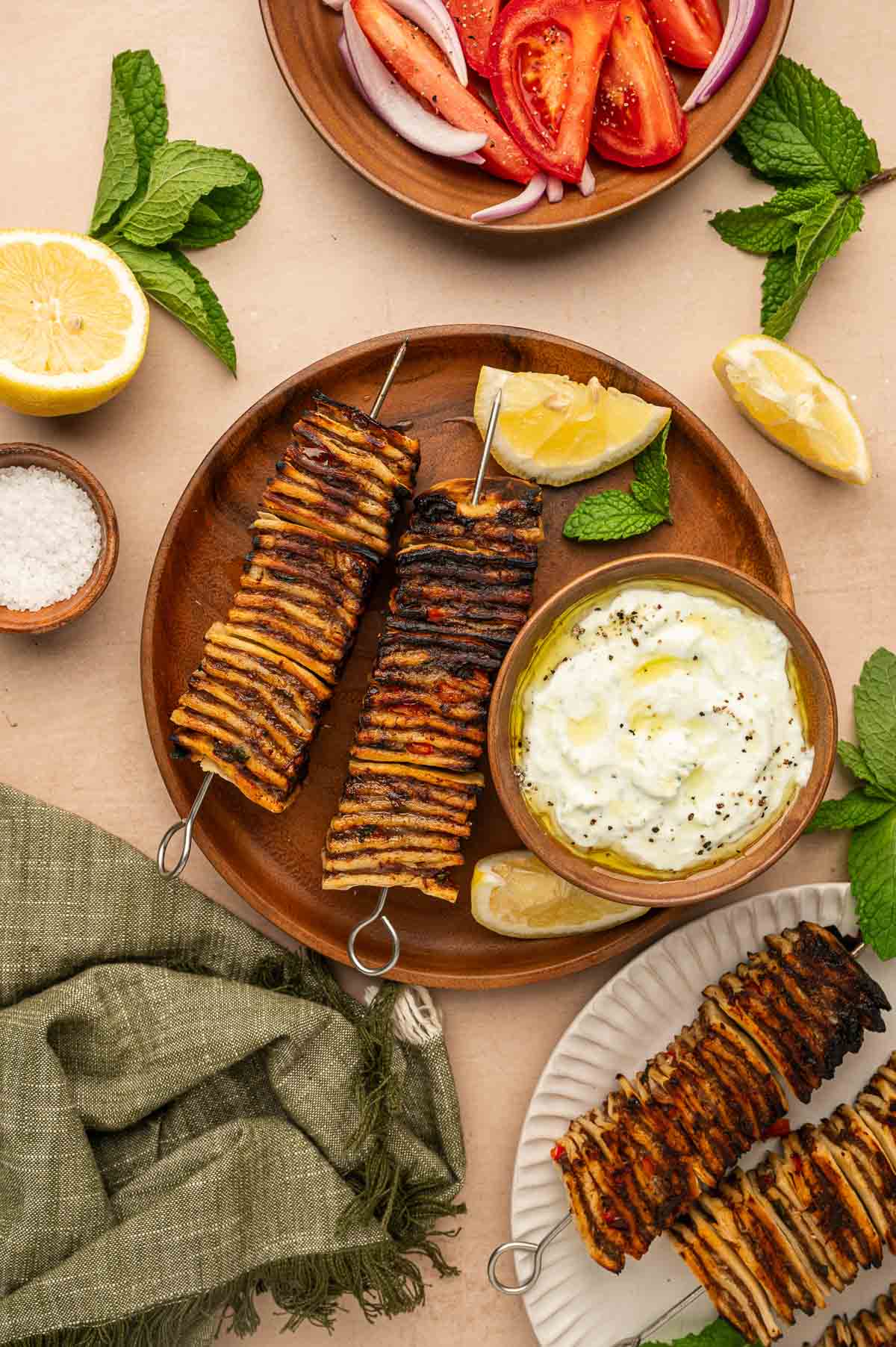 Grilled tortilla kebobs skewers on a plate with lemon wedges, a bowl of tzatziki, and a side of sliced tomatoes and onions. Mint leaves and a bowl of salt are also visible.
