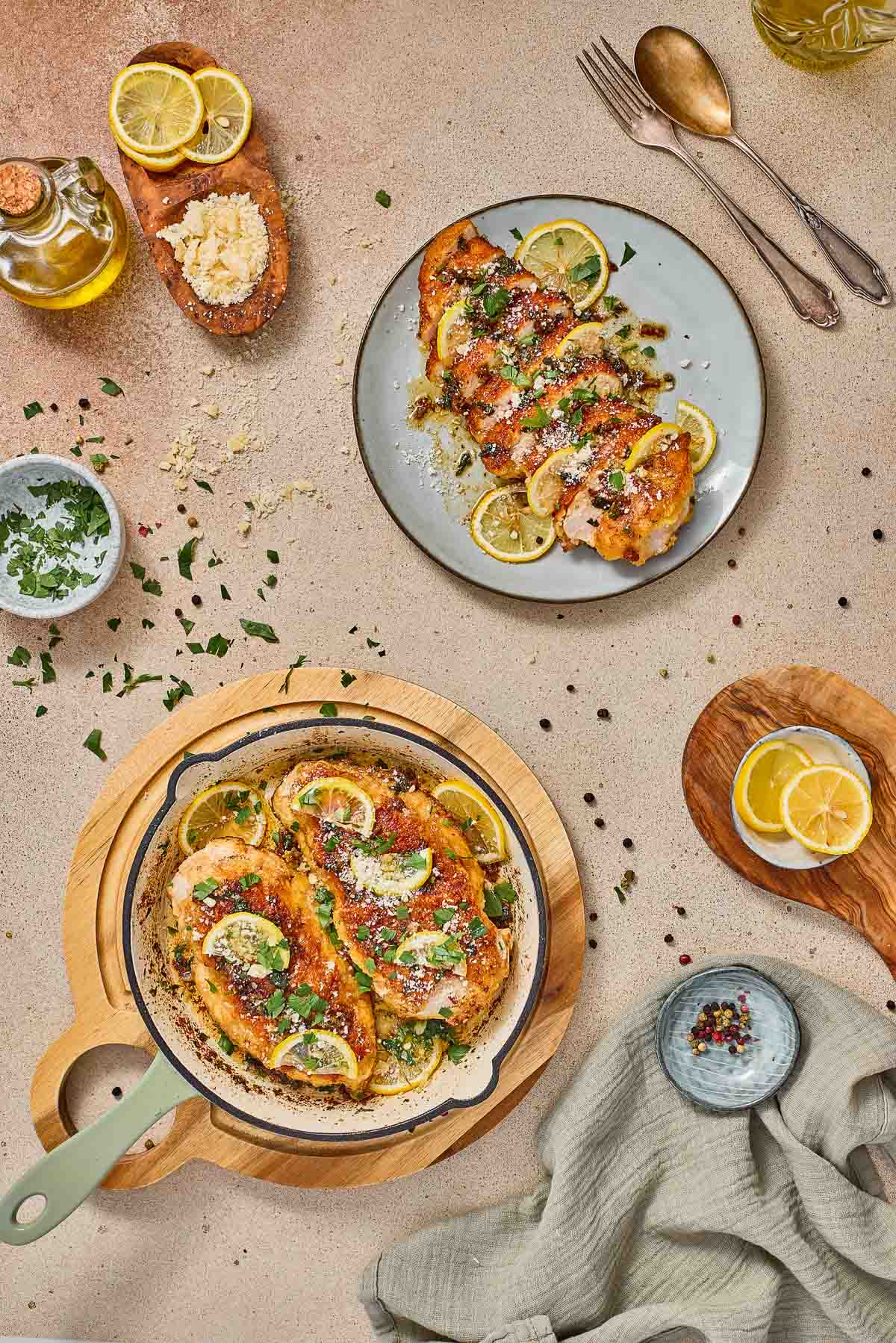 A top-down view of lemon chicken romano in a skillet, with a served portion on a plate, surrounded by lemon slices, parmesan, herbs, and kitchen utensils.