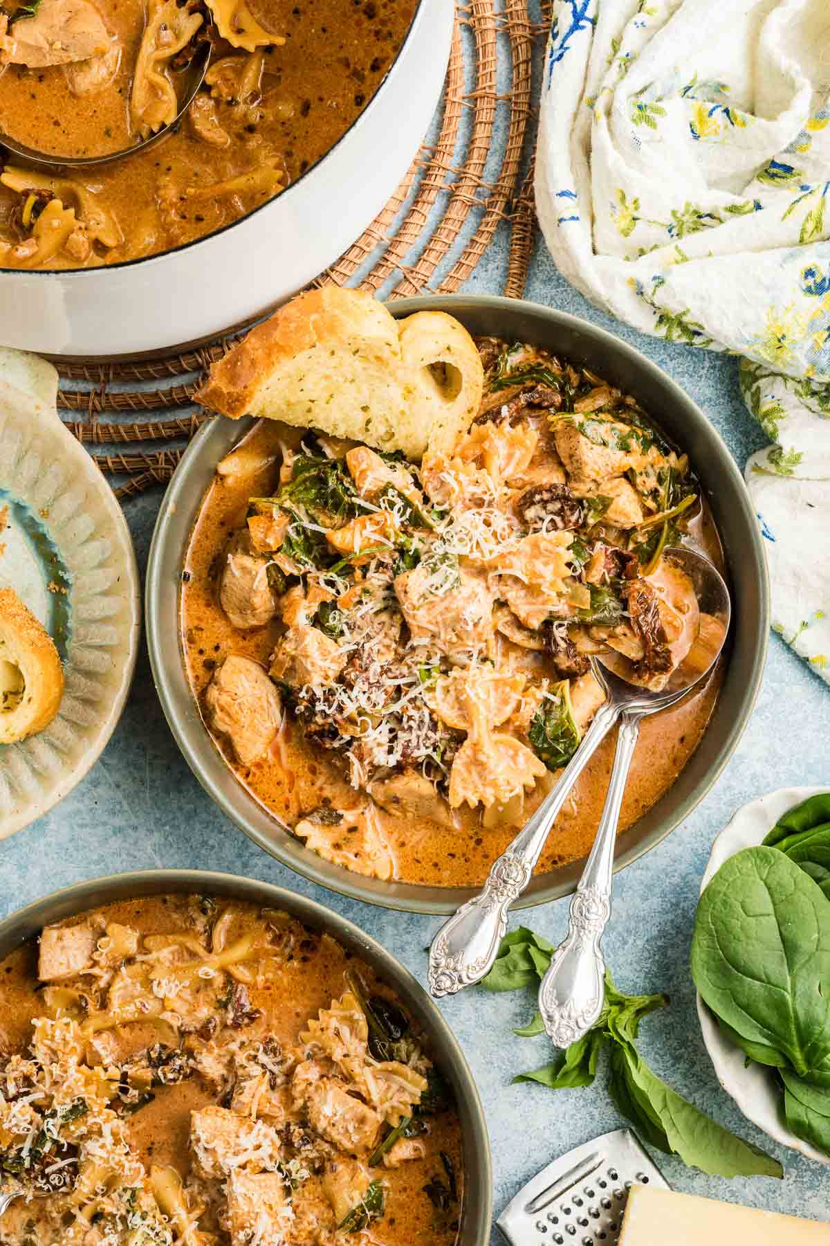 A bowl of creamy marry me chicken soup with pasta, spinach, chicken, and grated cheese, served with a slice of bread and two spoons. Other bowls and soup pot are visible nearby.