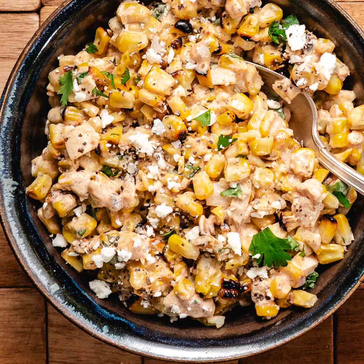 Mexican street corn salad with grilled corn, creamy dressing, cotija cheese, and fresh cilantro served in a black bowl with a serving spoon.
