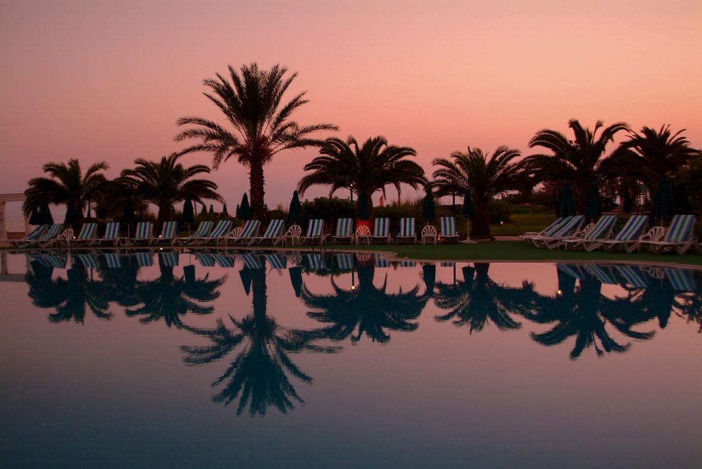 Resort swimming pool at dusk with palm trees and striped loungers