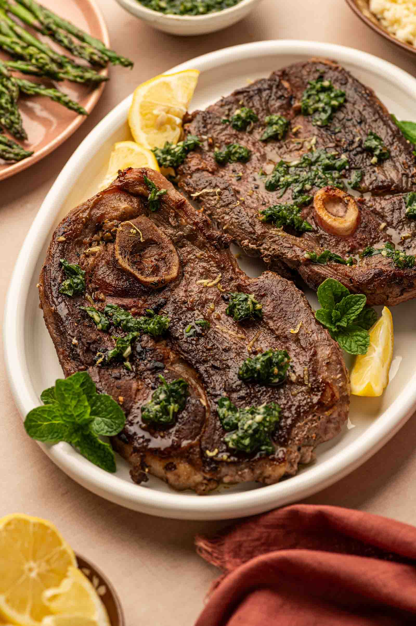 Two cooked lamb leg steaks with mint sauce, garnished with lemon wedges and mint, served on a white oval plate. Grilled asparagus and sauce bowls are in the background.