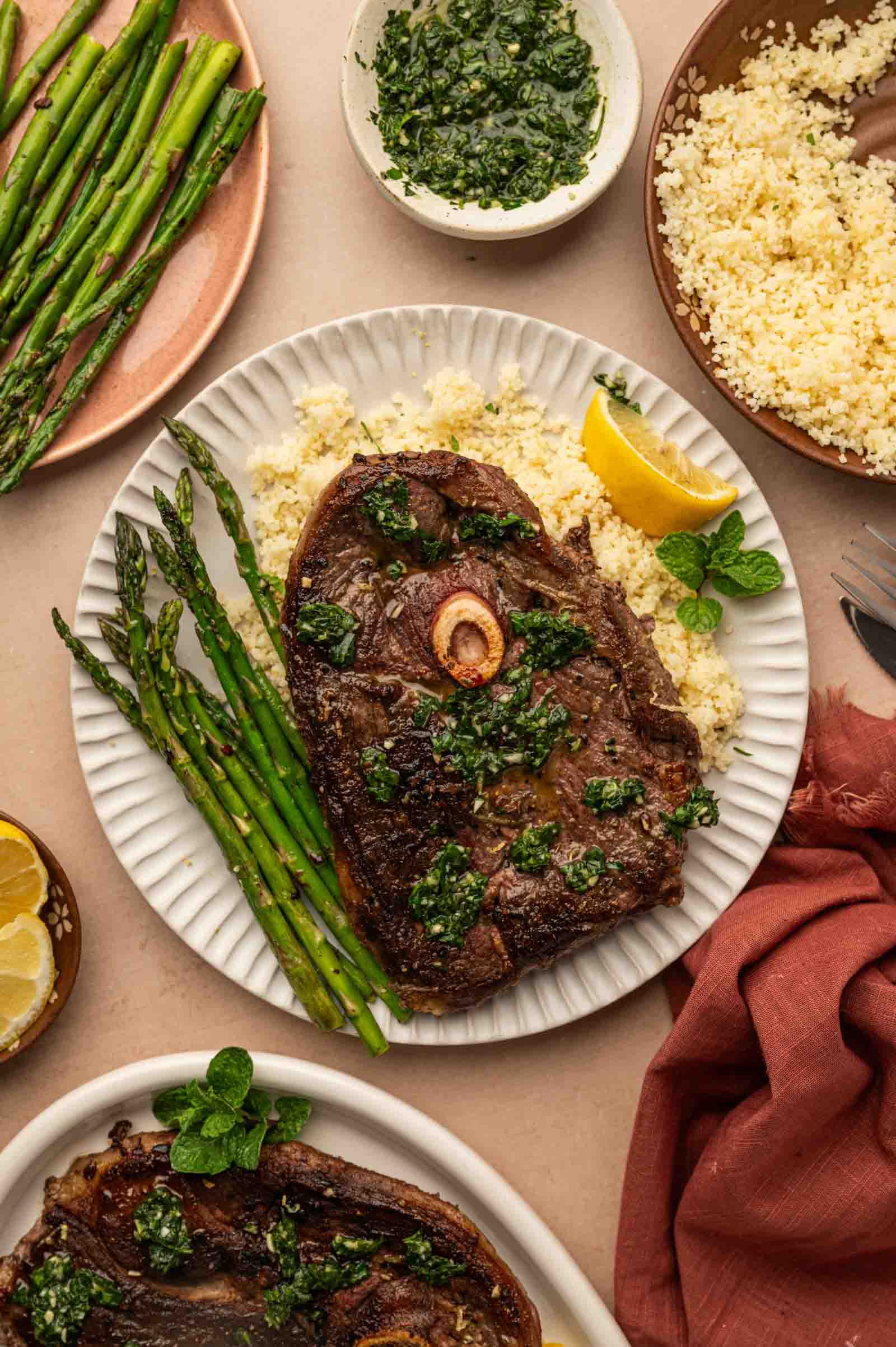 A plate with pan seared lamb steak topped with green sauce, served with asparagus, couscous, a lemon wedge, and garnished with mint; additional plates and bowls surround it.