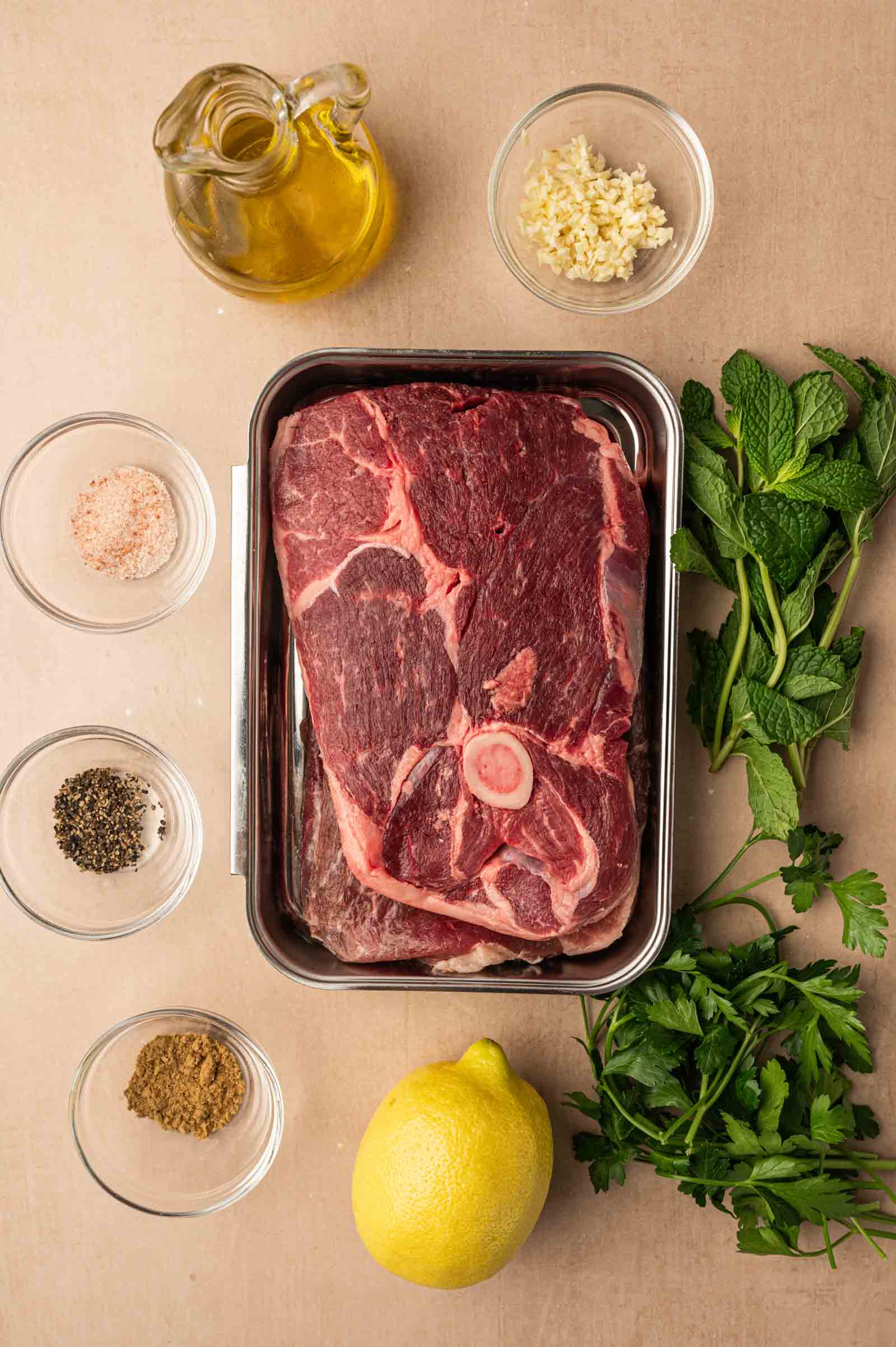 Raw lamb leg steaks in a metal tray surrounded by olive oil, chopped garlic, fresh mint, parsley, a lemon, ground spices, salt, and pepper in small bowls on a beige surface.