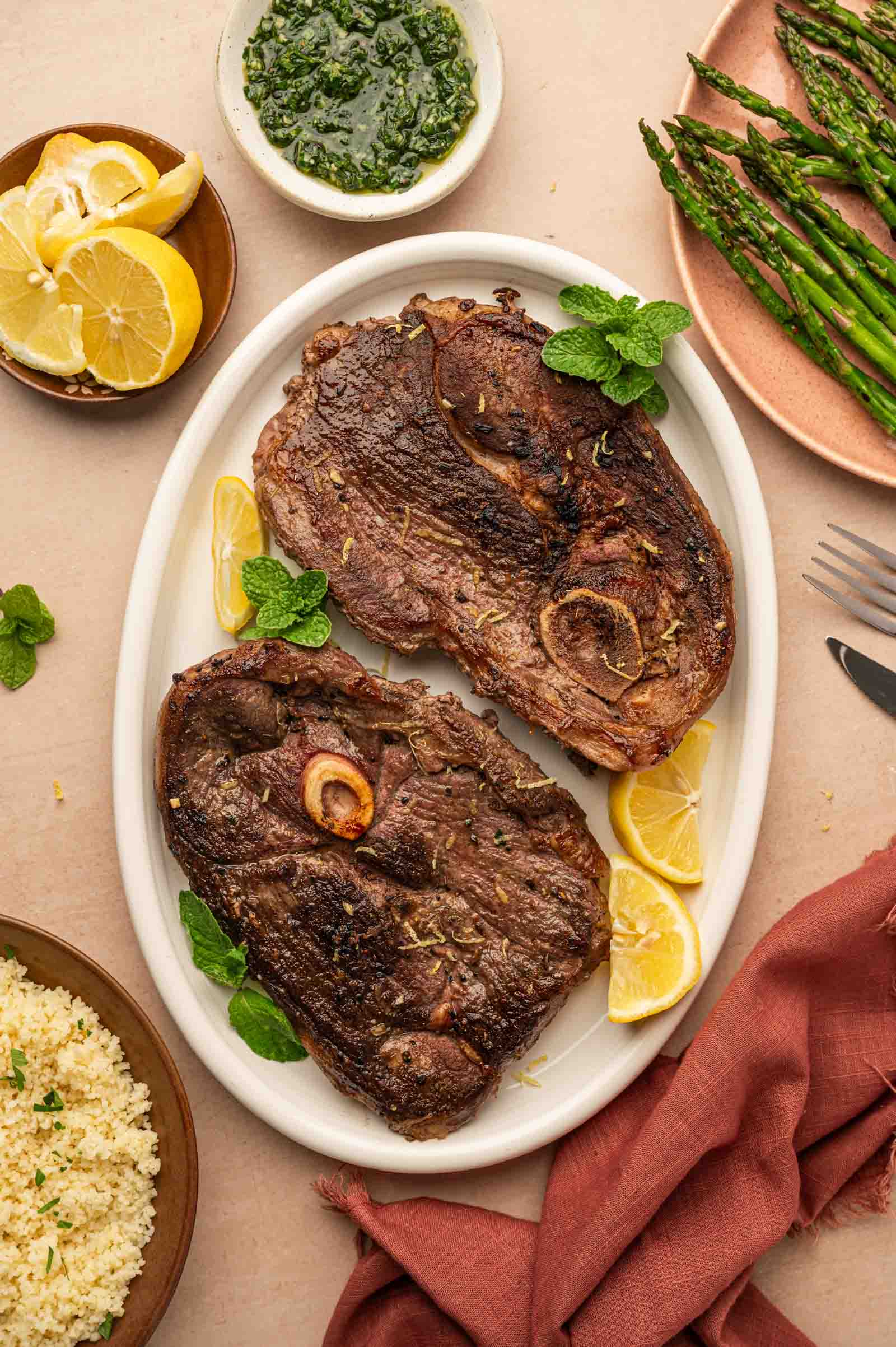 Two pan seared lamb leg steaks on a white oval plate with lemon wedges and mint leaves, surrounded by asparagus, couscous, lemon slices, and a bowl of green sauce.