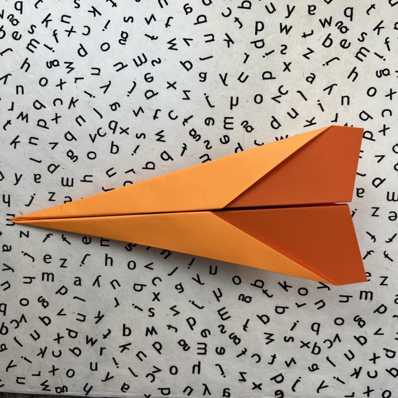 Finished Dart Plane Finished orange dart paper airplane on black and white alphabet background.