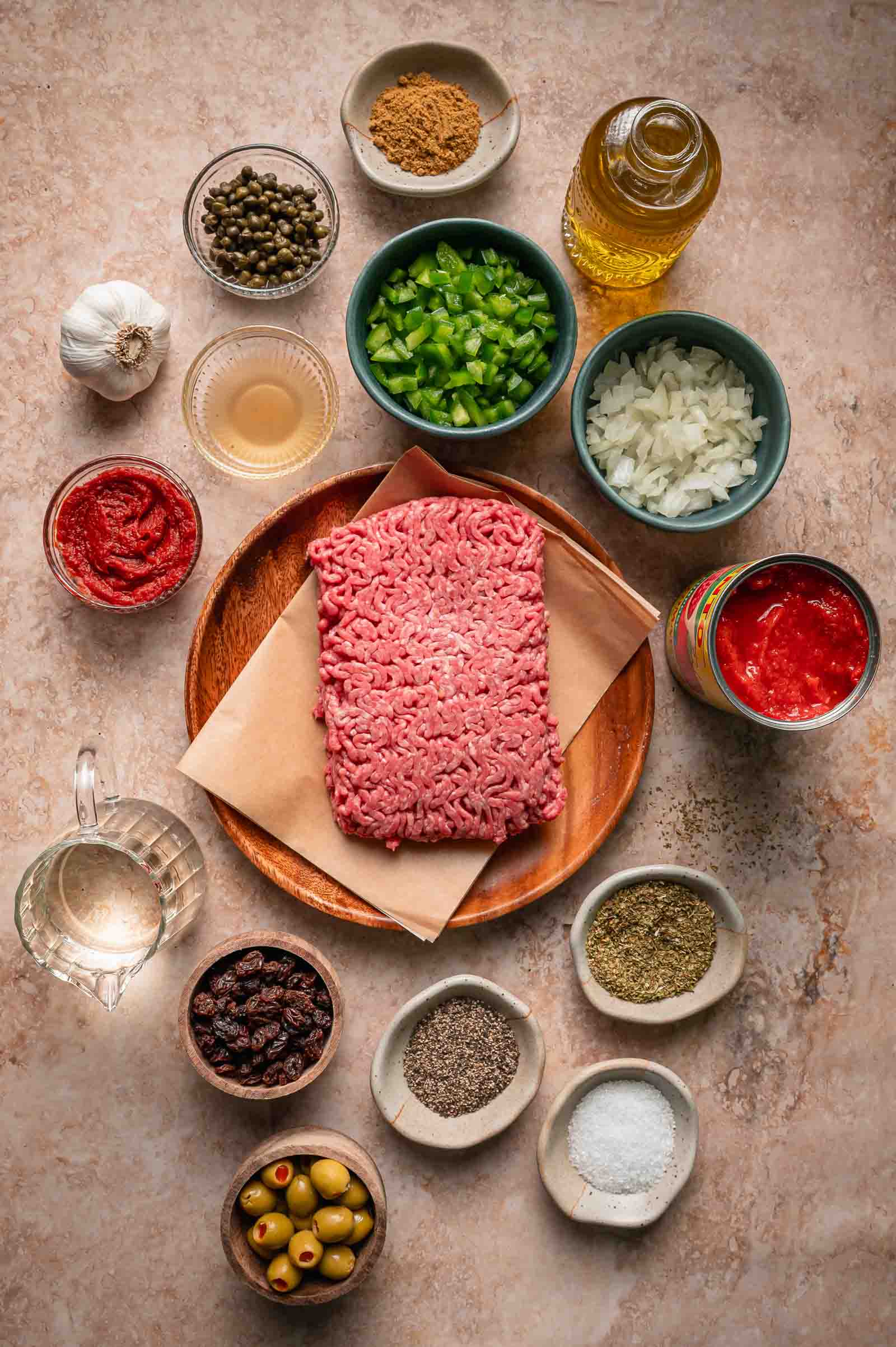 Raw ground beef on a wooden plate surrounded by small bowls of chopped vegetables, spices, olives, raisins, tomato products, garlic, and oil.