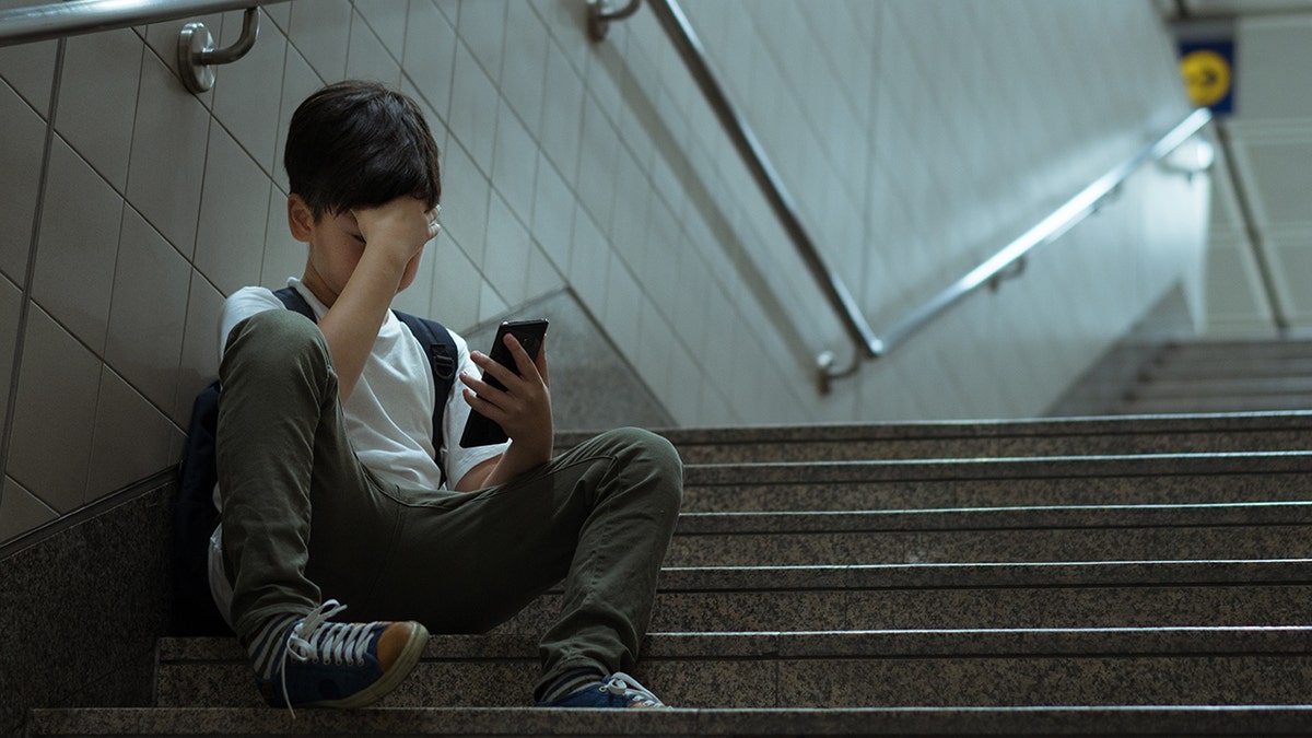 A kid covers his face while holding a phone and sitting on a staircase.