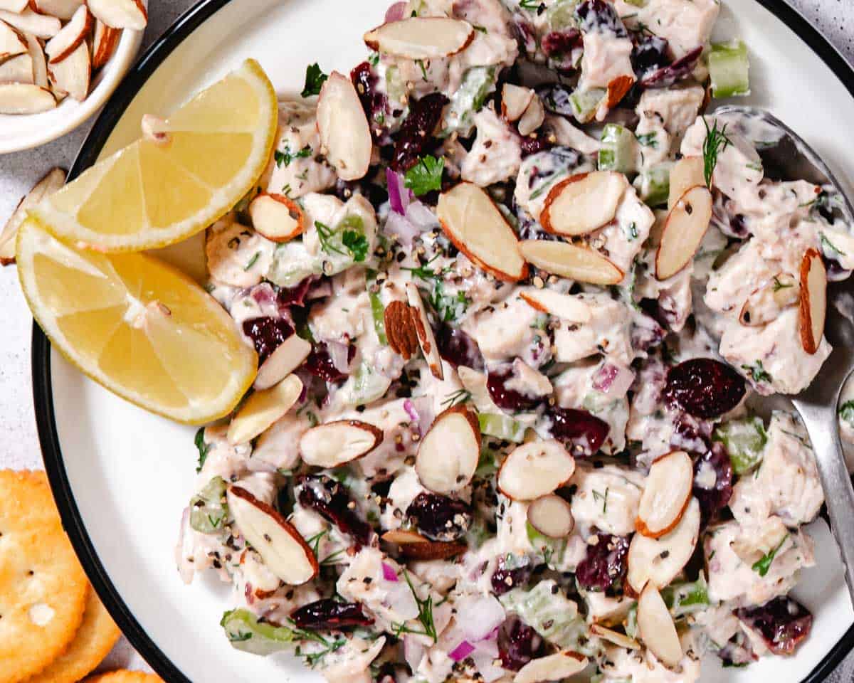 Creamy turkey salad with dried cranberries, celery, red onion, herbs, and sliced almonds on a white plate with lemon wedges.