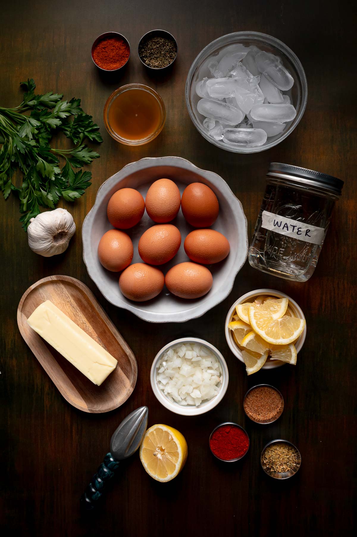 A flat lay of eggs, butter, chopped onion, garlic, parsley, spices, sliced lemon, ice water, and a citrus juicer arranged on a dark wooden surface.