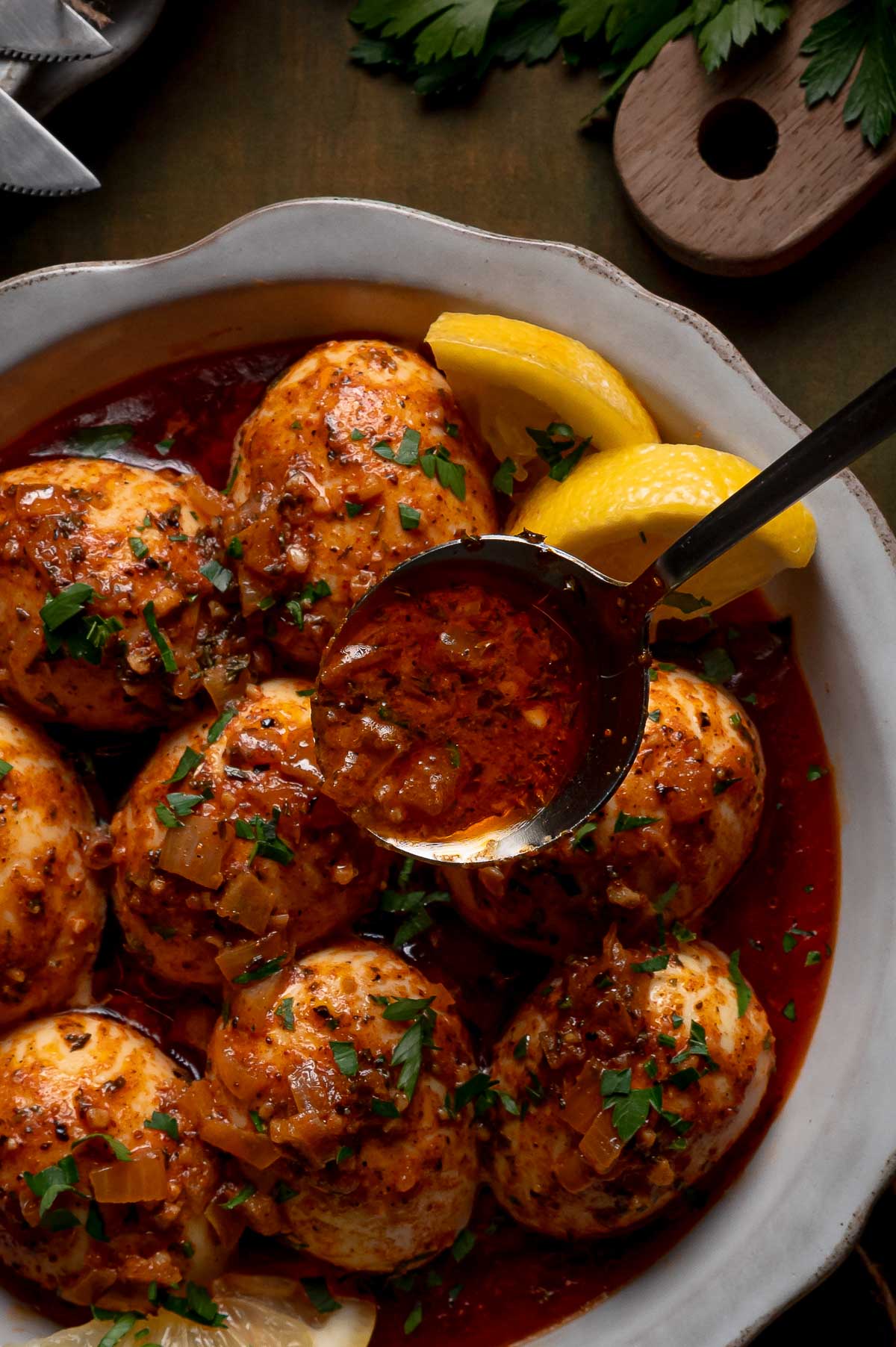 A bowl of hard-boiled eggs in a rich red spicy butter sauce, garnished with chopped herbs and served with lemon wedges.