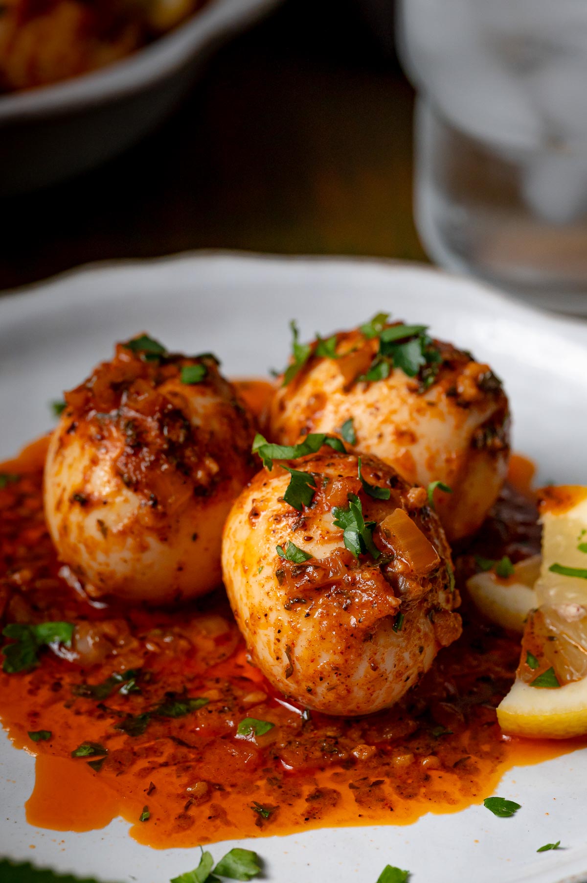 Three hard-boiled eggs covered in spicy red sauce, garnished with chopped herbs, served on a white plate with a lemon wedge.