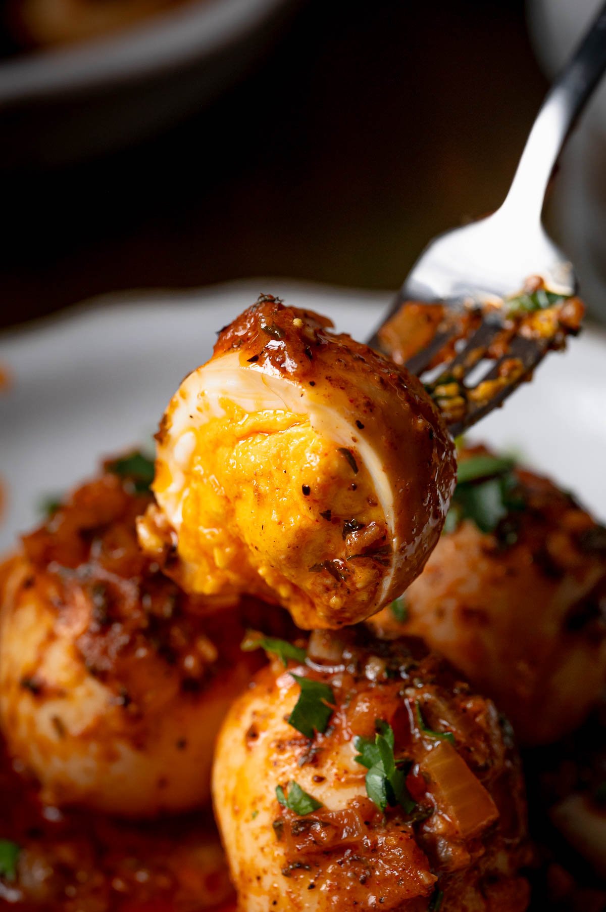 A fork holds a halved boiled egg coated in spicy red sauce, with other sauced eggs and chopped herbs on a plate below.