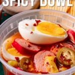 A plastic bowl filled with a spicy mixture, including a halved boiled egg, sausage slices, jalapeños, and red pepper flakes, with text about a viral spicy bowl recipe.