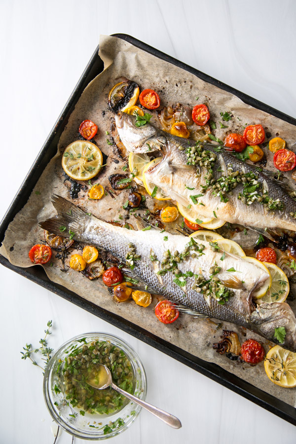 A baking tray with two whole roasted fish garnished with herbs, lemon slices, and surrounded by roasted cherry tomatoes. A glass bowl of green herb sauce with a spoon is placed nearby on a white surface.