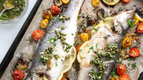 Two whole roasted fish on a baking sheet, garnished with lemon slices, capers, fresh herbs, and surrounded by roasted cherry tomatoes and garlic cloves. A bowl of herb dressing sits nearby.