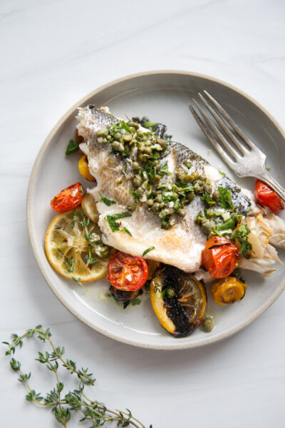 A plate of cooked white fish topped with chopped herbs and capers, served with roasted cherry tomatoes and lemon slices, garnished with fresh thyme, with a fork on the side.