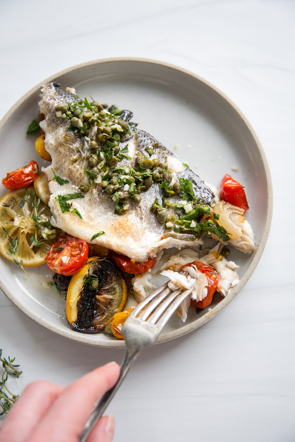 A plate with baked white fish topped with capers and herbs, served with roasted tomatoes and lemon slices; a hand holds a fork taking a bite from the fish.