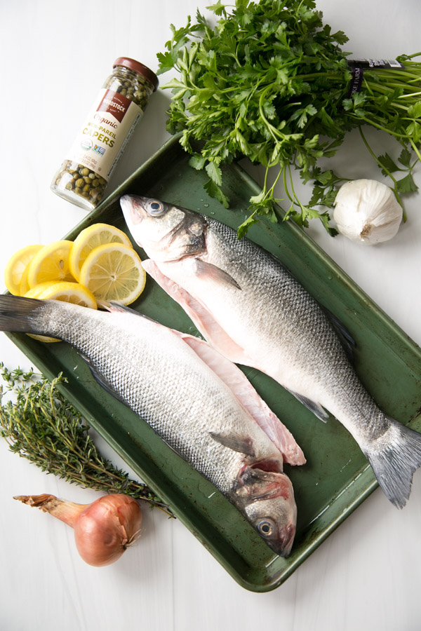 Two whole raw fish on a green baking tray, surrounded by fresh parsley, thyme, lemon slices, a shallot, garlic, and a jar of capers on a white surface.