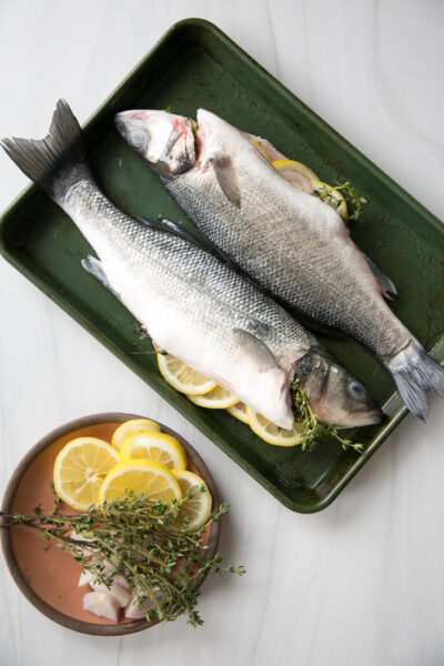 Two whole fish stuffed with lemon slices and fresh thyme are placed on a green baking tray. Nearby, a brown plate holds more lemon slices, thyme sprigs, and a few pieces of fish on a light surface.