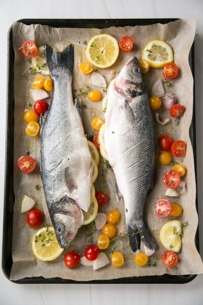 Two whole raw fish with lemon slices, cherry tomatoes, onions, and herbs are arranged on a baking sheet lined with parchment paper, ready to be roasted.