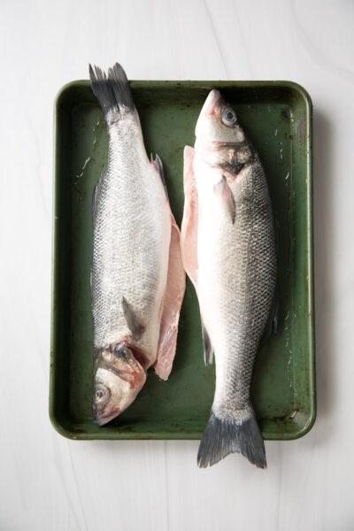 Two whole raw fish with silvery scales lie side by side on a green metal tray; one is partially filleted, exposing pink flesh. The tray rests on a white surface.