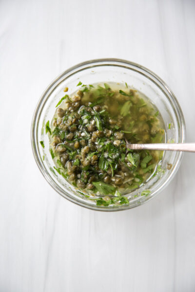 A glass bowl filled with a chopped green herb, caper, and olive oil mixture sits on a white surface with a spoon inside.