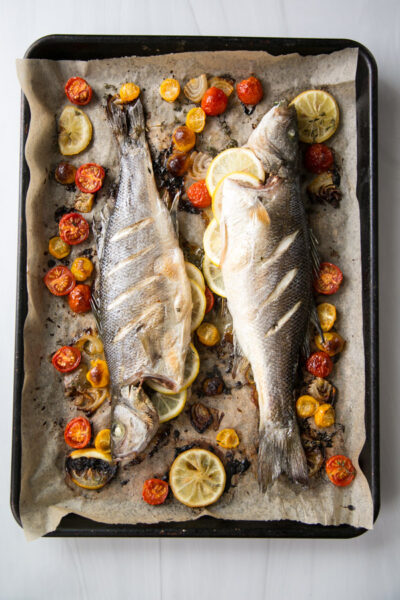 Two whole baked fish on a parchment-lined baking tray, surrounded by sliced lemons, cherry tomatoes, and onions. The fish have slits on their skin and are roasted with herbs and vegetables.