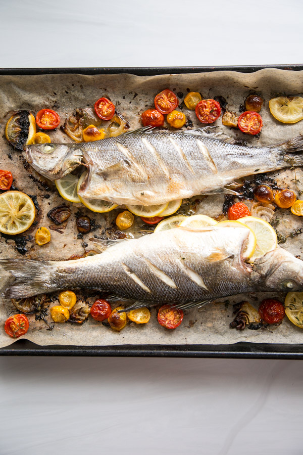 Two whole baked fish are placed on a baking tray lined with parchment paper, surrounded by roasted cherry tomatoes and lemon slices. The fish have scored skin and are garnished with lemon.