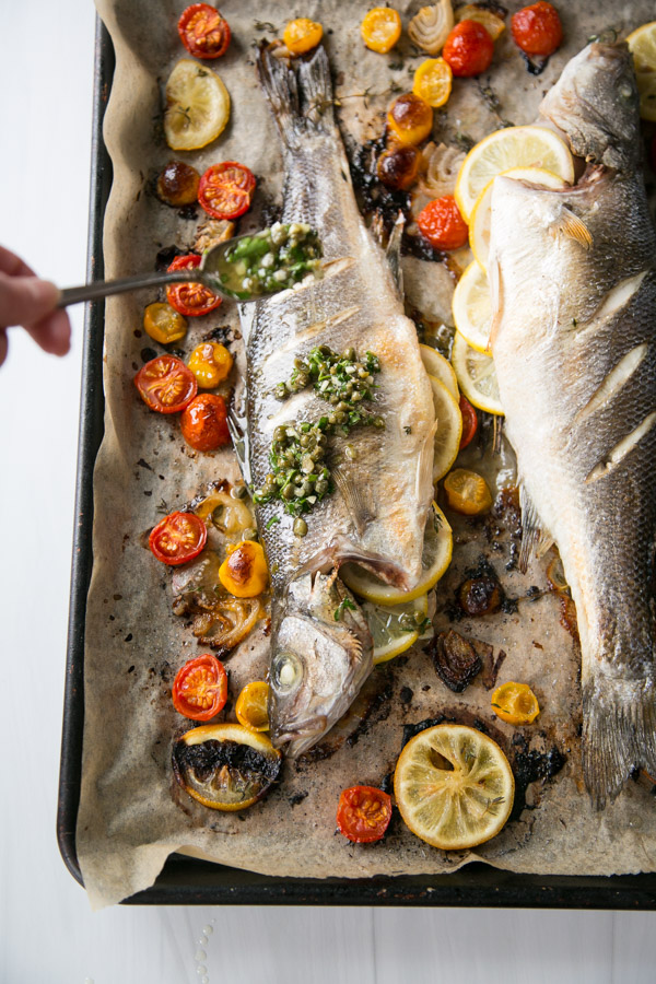 Two whole baked fish on a parchment-lined tray, garnished with sliced lemons, roasted cherry tomatoes, and herbs. A hand is spooning a green sauce onto one of the fish.