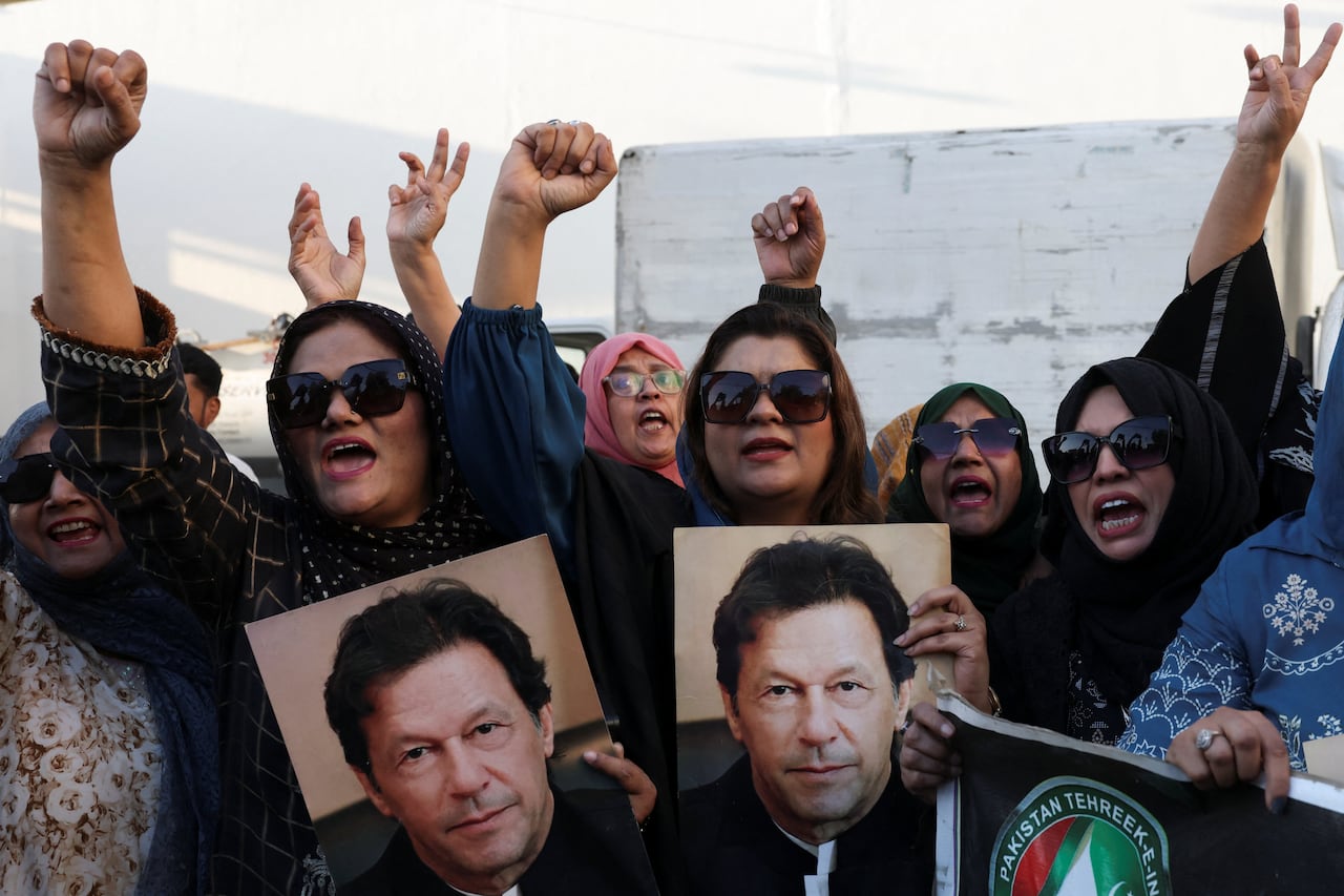 A crowd of protesters stands outside. Three veiled women in sunglasses are at the front, pumping their fists and holding photos of a black-haired man.