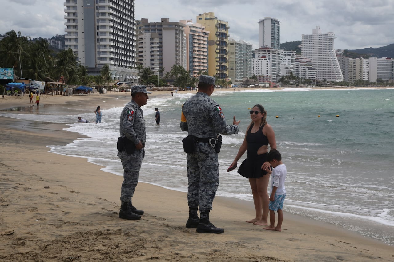 Two soldiers in uniform are shown on a beach talking to a woman who is standing with a small boy. In the background, other beachgoers and hirise buildings are shown.
