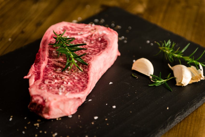 Waygu New York strip steak on a slate board with flakey salt, garlic cloves and rosemary.