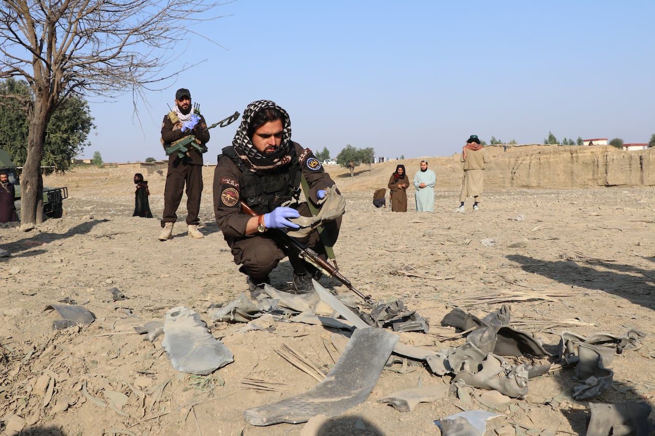 A bearded man in uniform wearing a hood crouches over pieces of metal debris on the dirt ground.