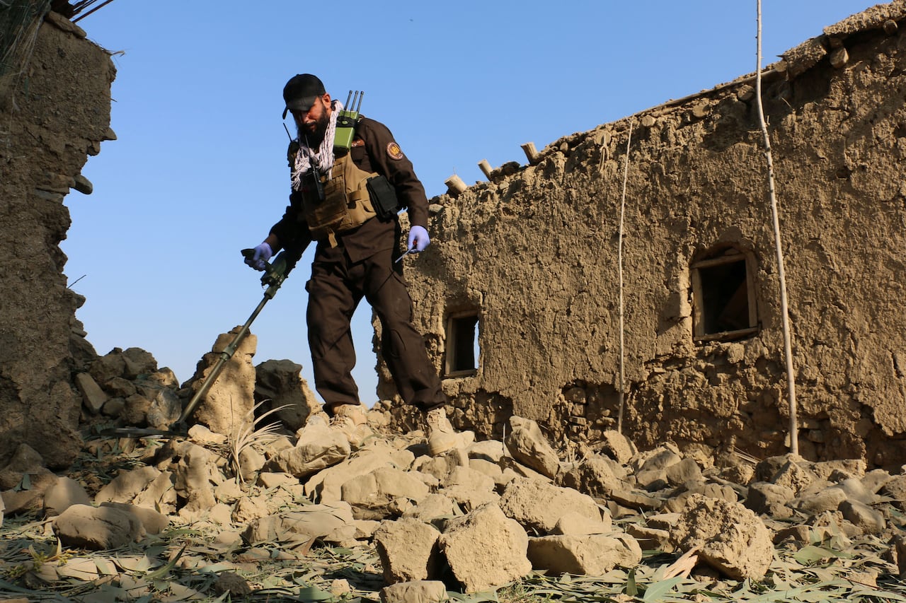 A bearded man in a uniform with a metal detector stands near piles of rubble between buildings on the ground.