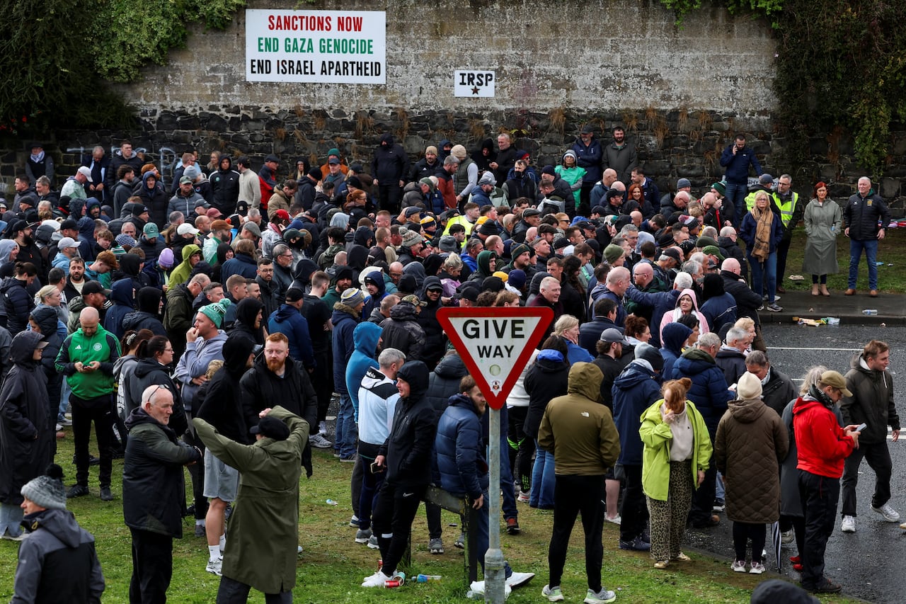 A group of people gather on a patch of grass with a wall with a sign saying "Sanctions now; end Gaza genocide; end Israel apartheid."