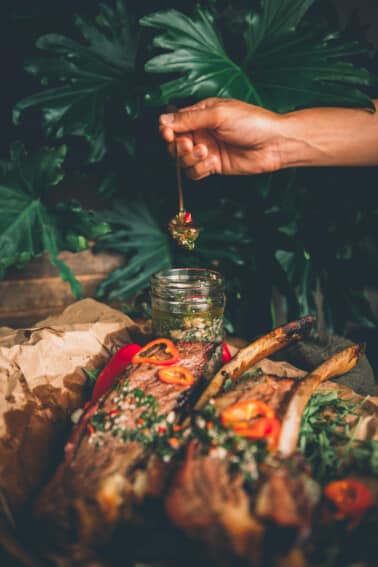 Hand holding spoon full of chimichurri over a jar with beef ribs slathered in the sauce in the foreground.
