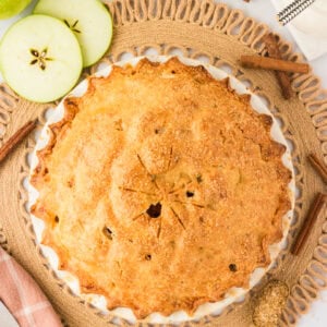 A golden-brown gluten-free apple pie with a flaky crust sits in a white pie dish, sprinkled with coarse sugar. The pie is surrounded by sliced green apples, cinnamon sticks, and a rustic woven placemat.